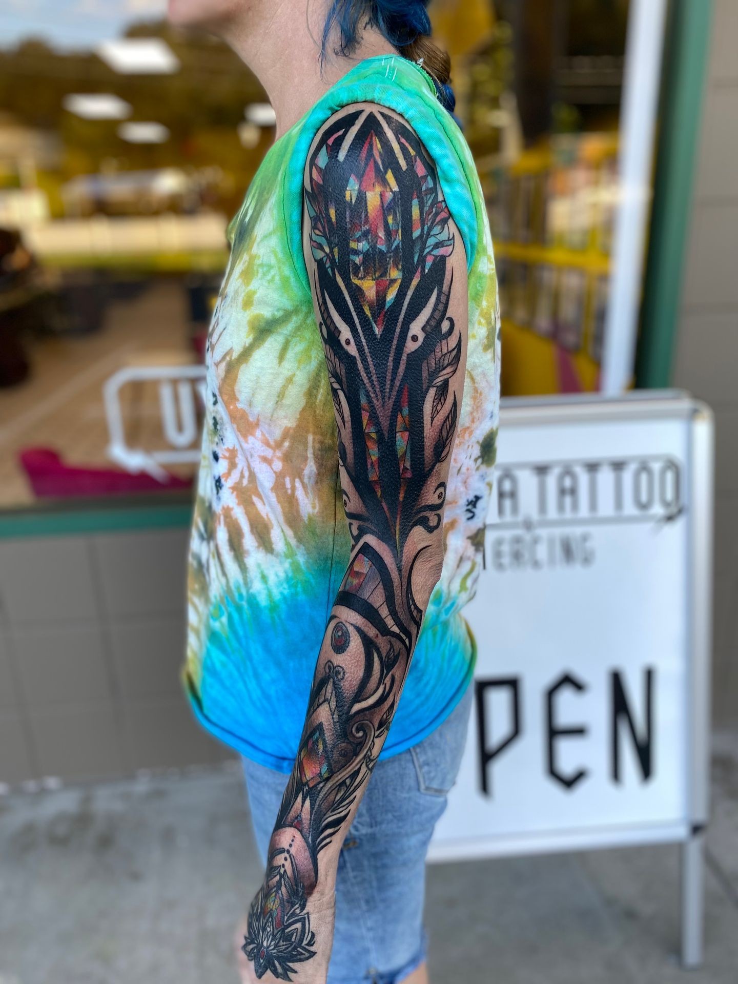 Person with colorful geometric tattoo sleeve wearing a tie-dye shirt and denim shorts.