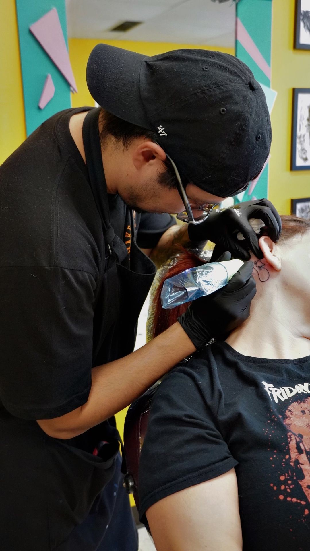 Tattoo artist with black gloves creating a tattoo behind a person's ear in a brightly colored room.