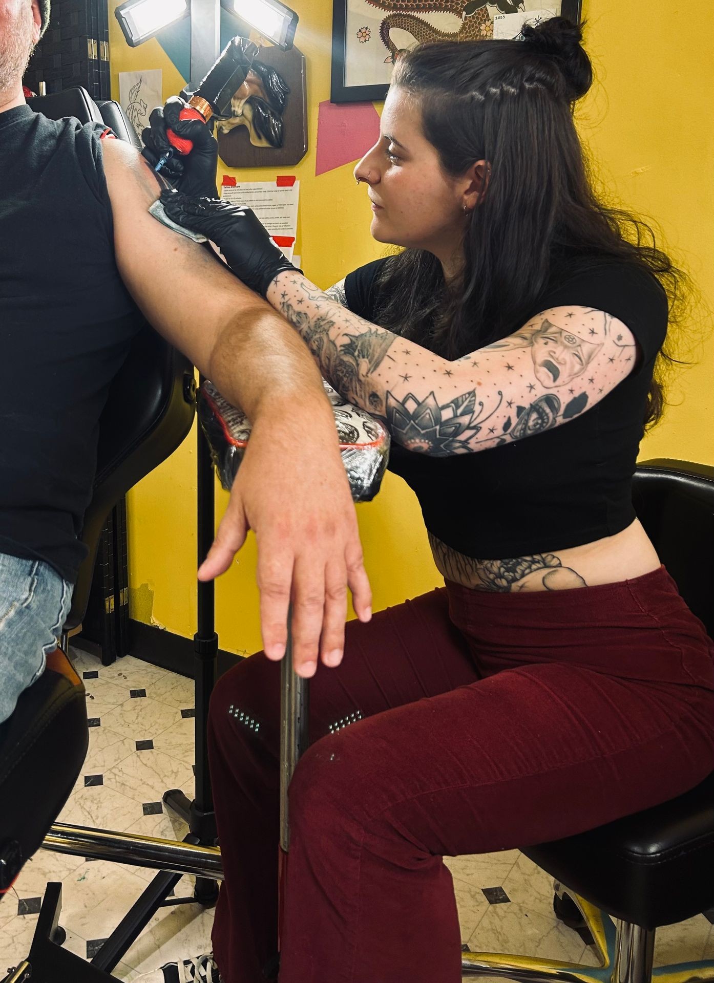 Tattoo artist with tattooed arm working on a client's arm in a brightly lit studio.