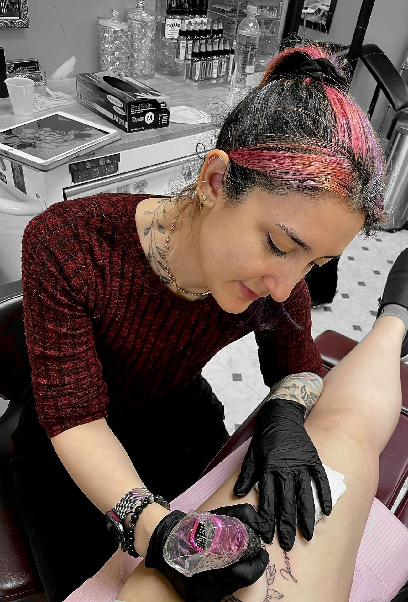 Tattoo artist in a red shirt with black gloves working on a tattoo on a client's leg.