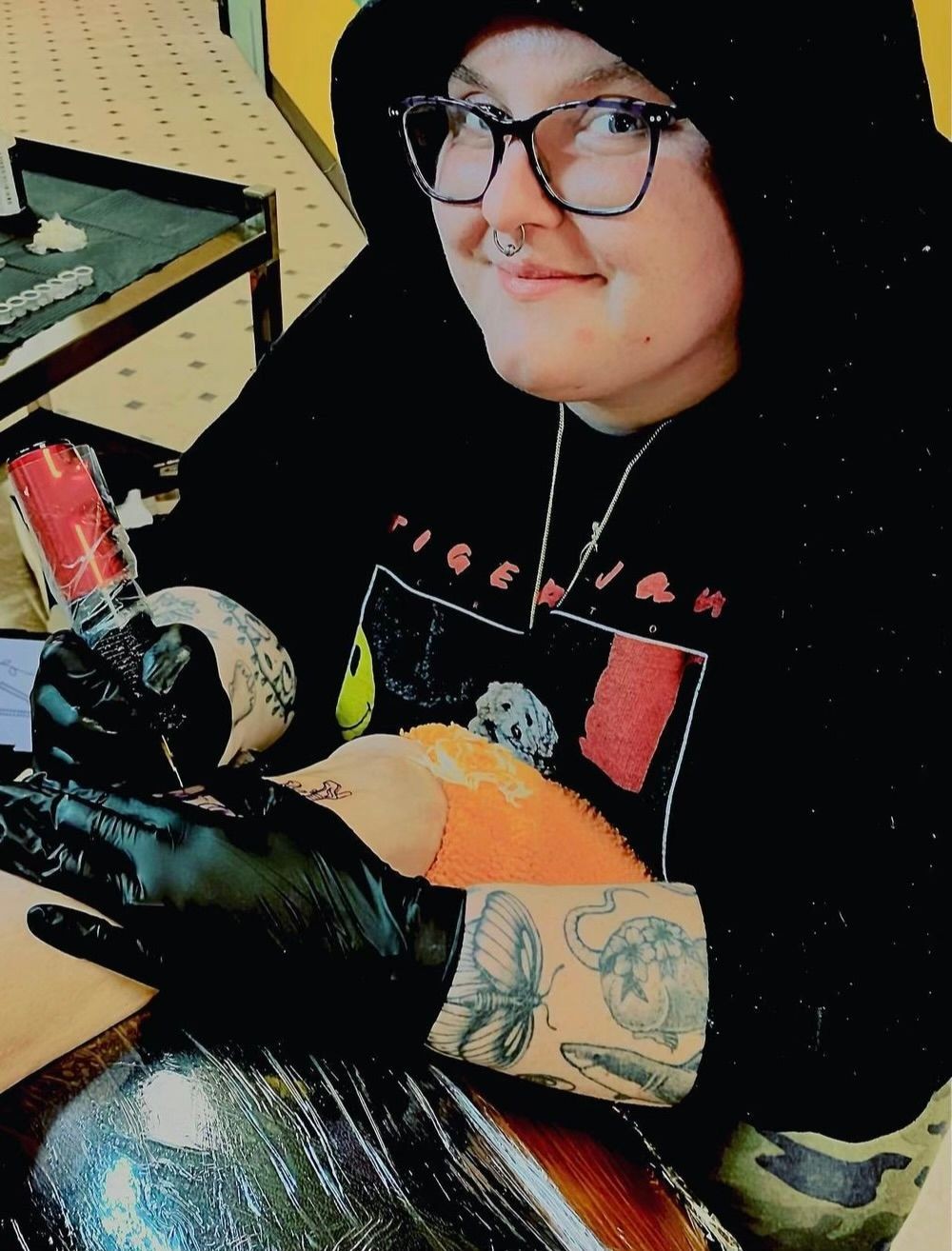 Person with tattoos and piercings giving a tattoo, wearing a black hoodie and gloves.